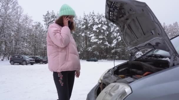 En kvinna ringer på sin smartphone när hon står vid trasig bil med öppen bilhuva på vintern. — Stockvideo