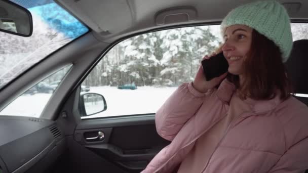 Menina bonita nova em uma jaqueta rosa para baixo e chapéu de inverno falando use seu smartphone enquanto está sentado no banco do passageiro de um carro — Vídeo de Stock