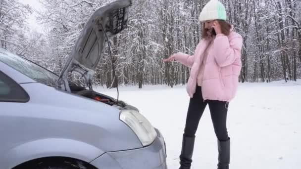 Μια γυναίκα καλεί στο smartphone της, ενώ στέκεται δίπλα στο σπασμένο αυτοκίνητο με ανοιχτή κουκούλα αυτοκινήτου το χειμώνα. — Αρχείο Βίντεο