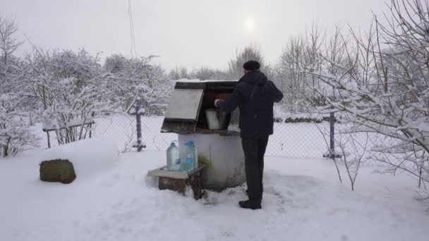 Человек берет ведро воды из колодца и выливает его в пластиковые бутылки во время снегопада. — стоковое видео