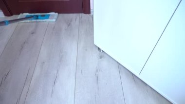 Kapının yakınını temizliyorum. Dolabın altını temizliyorum. Mavi paspasla gri ahşap zemini temizliyorum. Yakın plan.
