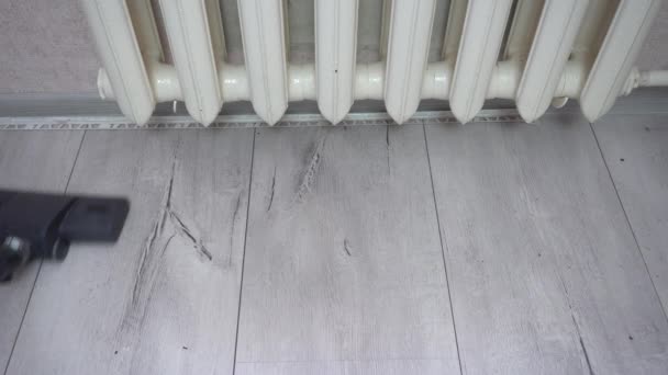 El cepillo de aspiradoras absorbe el polvo en la superficie laminada gris. El cepillo de aspiradora limpia el suelo de madera gris cerca del radiador del calentador. Limpieza general semanal de la criada — Vídeos de Stock