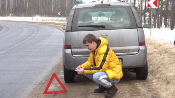 Le gars est assis près de la voiture cassée et cherche quelque chose au téléphone, le gars ne sait pas quoi faire et la voiture a des feux de détresse allumés — Video