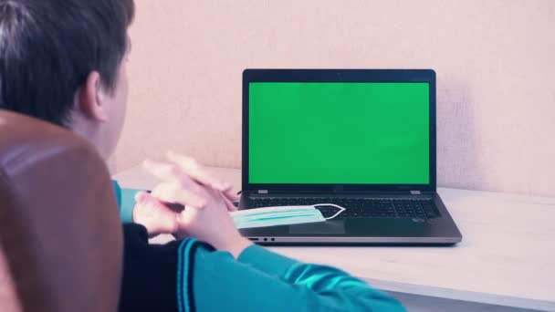 Un hombre se pone una mascarilla protectora antes de comenzar a trabajar en un ordenador portátil a través de la comunicación de vídeo, croma clave en la pantalla del ordenador portátil — Vídeos de Stock