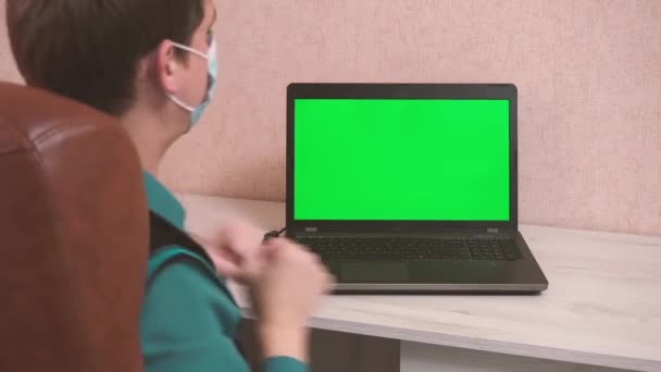 Un hombre baila en una computadora en una videoconferencia con una máscara protectora, una tecla de croma en la pantalla de un portátil y una alegría de hombre — Vídeos de Stock