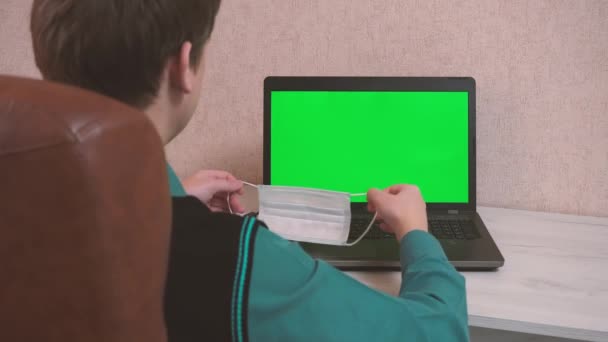 Muž si sám nasadí ochrannou masku obličeje a pomocí videohovoru naváže kontakt a začne psát na notebook, epidemiologickou bezpečnost, dálkovou práci, zelenou obrazovku — Stock video