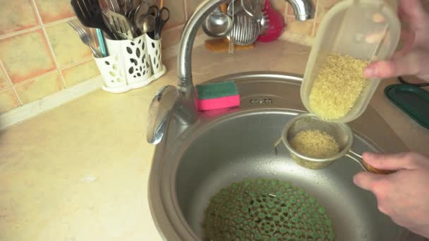 A portion of rice is poured into a colander and washed under running water in the sink — Wideo stockowe