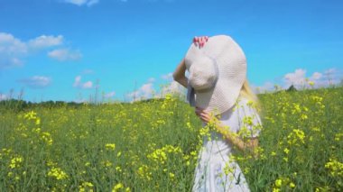 Genç ve güzel bir kız hasır bir şapkanın arkasına saklanıyor ve yazın açan bir kolza tarlasında eğlenmek için yüzünü buruşturuyor.