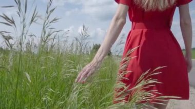 Kırmızı elbiseli genç bir kız, yazın yol kenarında yürüyor ve yeşil bitkilere eliyle dokunuyor. Kırmızı ojeli bir kadın, bitkilere dokunuyor.