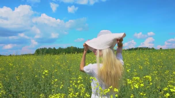 Молода красива дівчина в білій сукні одягає елегантний капелюх на голову, дівчина позує і посміхається в квітучому полі ріпаку влітку — стокове відео