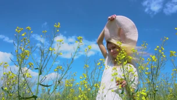 Jong mooi meisje in een witte jurk gooit uit haar brede hoed, het meisje poseert en glimlacht in een bloeiende koolzaad veld in de zomer, bottom view — Stockvideo