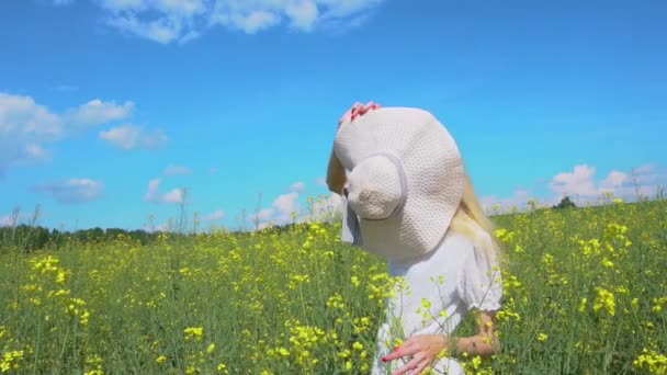 Молода красива дівчина ховається за солом'яним капелюхом, а потім показує своє обличчя посмішкою в квітучому полі ріпаку влітку — стокове відео