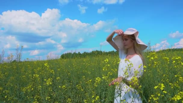 Молода красива дівчина в білій сукні відкидає свій широкий капелюх, дівчина позує і посміхається в квітучому полі ріпаку влітку — стокове відео