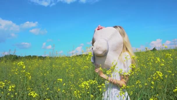 Молода красива дівчина ховається за солом'яним капелюхом, а потім показує своє сумне обличчя в квітучому полі ріпаку влітку — стокове відео