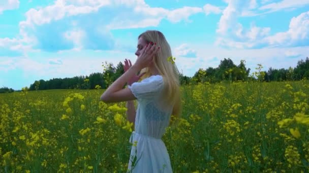 Gadis muda meluruskan rambutnya yang panjang di ladang jerami yang mekar, menyentuh bunga-bunga pemerkosa dengan tangannya pada hari musim panas yang cerah — Stok Video