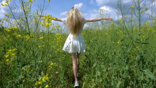 Tânără fată frumoasă într-o rochie albă se plimbă printr-un câmp înflorit de rapiță și își ridică mâinile bucurându-se de viață și libertate. — Videoclip de stoc