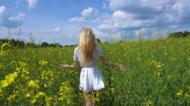 Una joven hermosa con un vestido blanco corre a través de un campo de colza floreciente balanceando su pelo largo y sonriendo en el verano — Vídeo de stock
