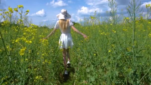 Молода красива дівчина в білій сукні в широкому капелюсі проходить квітучим ріпаковим полем, торкається рослин руками і піднімає руки, насолоджуючись життям і свободою — стокове відео