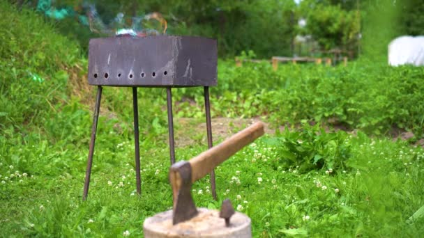 La parrilla casera oxidada de metal quema y fuma en el patio trasero en un día soleado y un hacha sobresale en la cubierta — Vídeo de stock