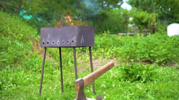 An einem sonnigen Tag brennt und raucht im Hinterhof ein rostiger hausgemachter Metallkohlegrill und eine Axt ragt aus dem Deck — Stockvideo
