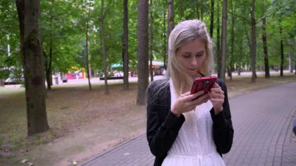 Menina bonita nova em um vestido branco e um casaco preto usa um smartphone no meio de uma passarela em um parque de diversões — Vídeo de Stock