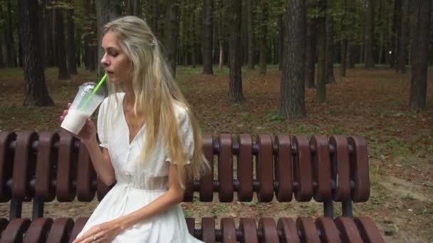 Egy fiatal, gyönyörű lány fehér ruhában ül egy padon a parkban, tejturmixot iszik, és aranyosan mosolyog érzelmileg. — Stock videók