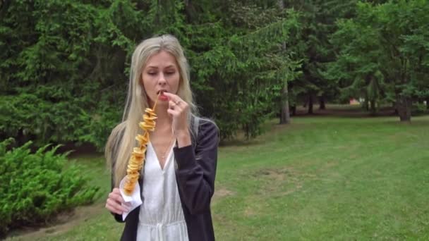 En ung vacker flicka med blont hår i en vit klänning och en svart kofta äter en gatumat tornado potatis i en nöjespark och berättar känslomässigt något och ler vackert — Stockvideo