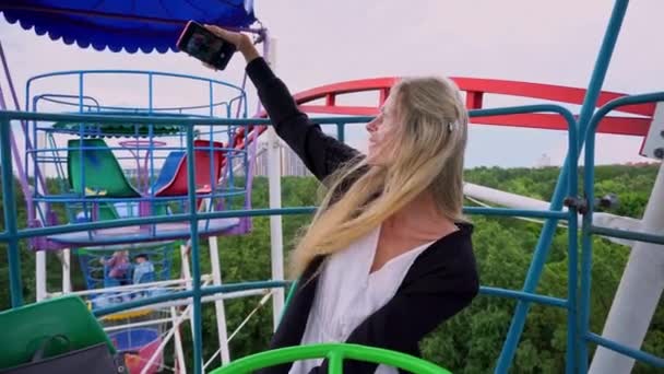 Fiatal gyönyörű lány fehér ruhában és egy fekete kardigán csinál szelfi okostelefonnal az óriáskeréken a vidámparkban — Stock videók