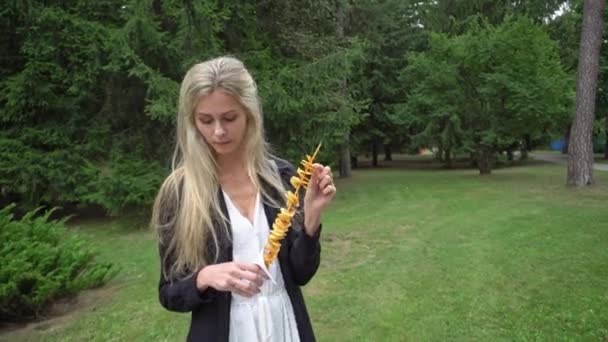 Una joven hermosa con el pelo rubio en un vestido blanco y un cárdigan negro come una comida callejera tornado-patatas en un parque de atracciones y emocionalmente dice algo y sonríe maravillosamente — Vídeo de stock