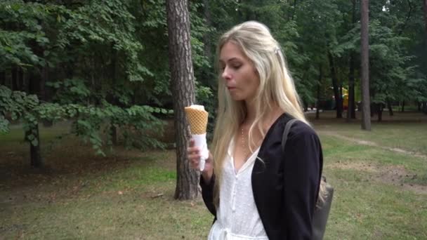 Seorang gadis muda cantik dalam gaun putih dan kardigan hitam di taman dan makan es krim wafel kerucut dan tersenyum cerdik sangat emosional — Stok Video