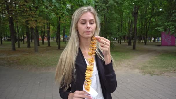 Een jong mooi meisje met blond haar in een witte jurk en een zwart vest eet een straat voedsel tornado-aardappelen in een pretpark en emotioneel vertelt iets en glimlacht prachtig — Stockvideo