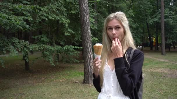 Een jong mooi meisje in een witte jurk en zwart vest in het park en eet een ijsje wafel kegel en glimlacht schattig heel emotioneel — Stockvideo