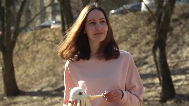 Une fille dans un pull rose regarde au loin et touche un jouet de poulpe tricoté sur un fond d'arbres d'automne — Video
