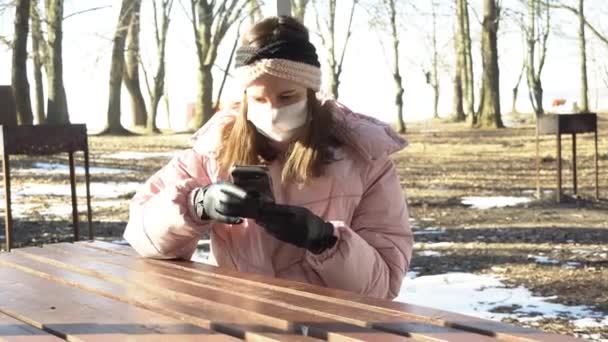 保護面マスクの女の子は、電話を使用し、公共の公園でのビデオ会議を介してコロナウイルスの中にデバイスを使用するために彼女の手袋を脱いで、コロナウイルスの制限 — ストック動画