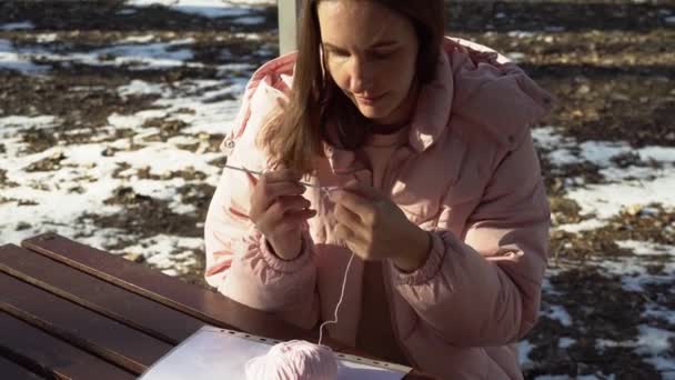 Κορίτσι πλέκει στο δρόμο σε ένα πάρκο της πόλης το χειμώνα στο ηλιοβασίλεμα, η διαδικασία του πλεξίματος στην ύπαιθρο — Αρχείο Βίντεο