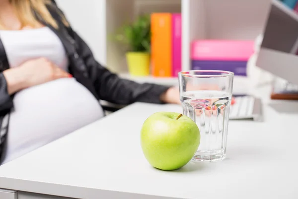 Kobieta w ciąży w biurze z jabłkiem i szkło wodne — Zdjęcie stockowe