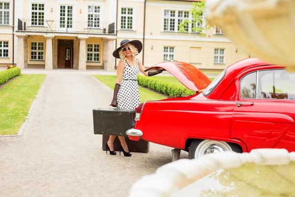 Женщина в кофейном платье с винтажным чемоданом рядом с красным ретро-автомобилем — стоковое фото