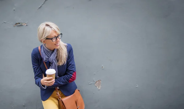 Μοντέρνα νεαρή γυναίκα στέκεται στον τοίχο σε εξωτερικούς χώρους και κρατώντας ένα φλιτζάνι καφέ. — Φωτογραφία Αρχείου