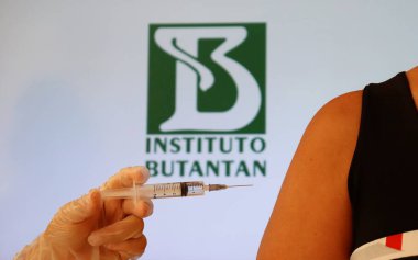 Bahia, Brazil - March 23, 2021. Hand holding a syringe against woman's shoulder, with Instituto Butantan logo displayed in the background. Brazilian CoronaVac Covid-19 vaccine. clipart
