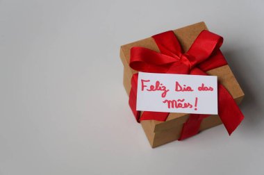 Kırmızı fiyonklu hediye kutusu ve Portekizce 'de' Anneler Günün kutlu olsun 'yazısı (Feliz Dia das Maes).