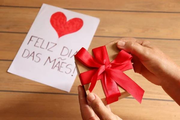 Mom holding gift box and 'Happy Mother's Day' gift card in portuguese (Feliz Dia das Maes) in the background.