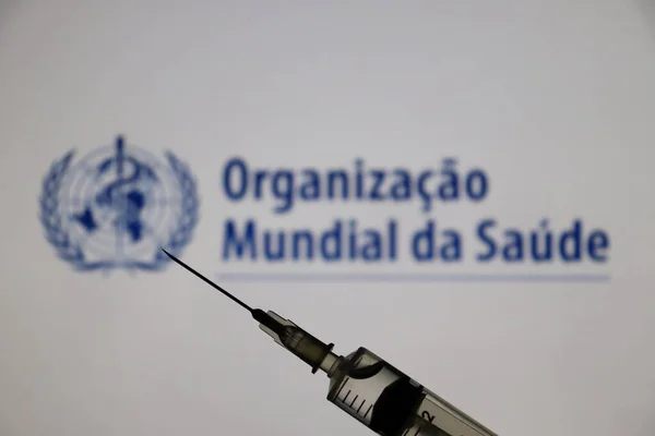 Bahia, Brezilya - 3 Mayıs 2021: Dünya Sağlık Örgütü (Organizacao Mundial da Saude - OMS, Portekizce) logosu ekranda ve elinde şırıngayla sergilenmektedir. Covid-19 aşı konsepti. 