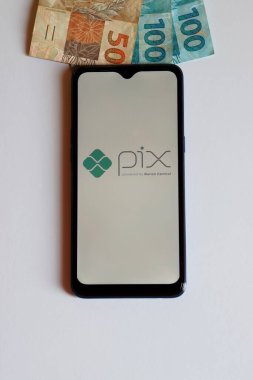Bahia, Brazil - October 9, 2020: Pix logo on smartphone screen and Brazilian Real banknotes. Pix is an electronic method to make payments and transfer money. Vertical photo.