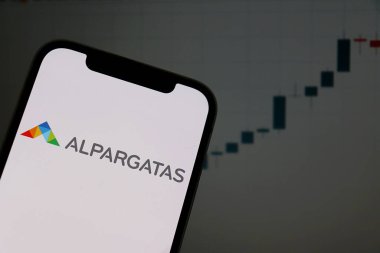 Salvador, Brezilya - 27 Aralık 2025: Akıllı telefon ekranında Alpargatas logosu ve arka planda borsa şamdan grafikleri. Alpargatas Brezilyalı şirket.