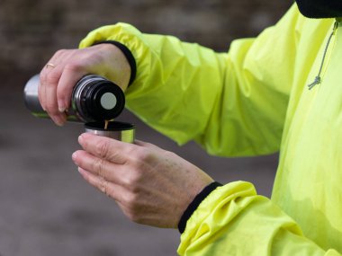Bir kadının kolları ve elleri metal bir gümüş şişeden şişeye sıcak bir içecek dökerken görülüyor. Kadın sarı bir hi-viz ceket giyiyor..