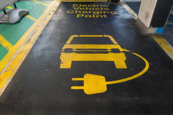 Otopark alanındaki çizgi işaretleri siyah arka planda sarı harflerle Elektrik Aracı Şarj Noktası diyor. Bir araba ve şarj kurşununun görüntüsü de görünür.