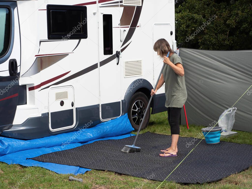 Al ady motorhome owner sweeps her outdoor mat with a broom outside her recreational vehicle. A bucket and wind break are visible