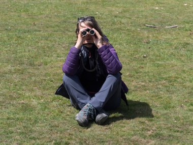 Bacak bacak üstüne atmış bir kadın dürbünle bakıyor. Yürüyüş botları ve giysileri giyiyor.