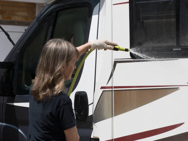 A lady motorhome owner cleans her recreational vehicle with a hose pipe.Spray can be seen on sunny day.