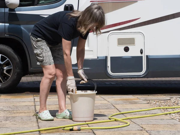 A lady motorhome owner has bucket, hose pipe ready to clean her recreational vehicle which can be seen in background.She wears shorts on a sunny day